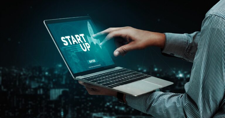 Pessoa segurando um laptop aberto enquanto toca a tela com uma das mãos. No display, aparece a palavra ‘STARTUP’ acompanhada da imagem de um foguete estilizado e um botão de ‘Enter’. Ao fundo, observa‑se uma cidade desfocada à noite, com diversas luzes urbanas criando um clima tecnológico e futurista. (sellentt)